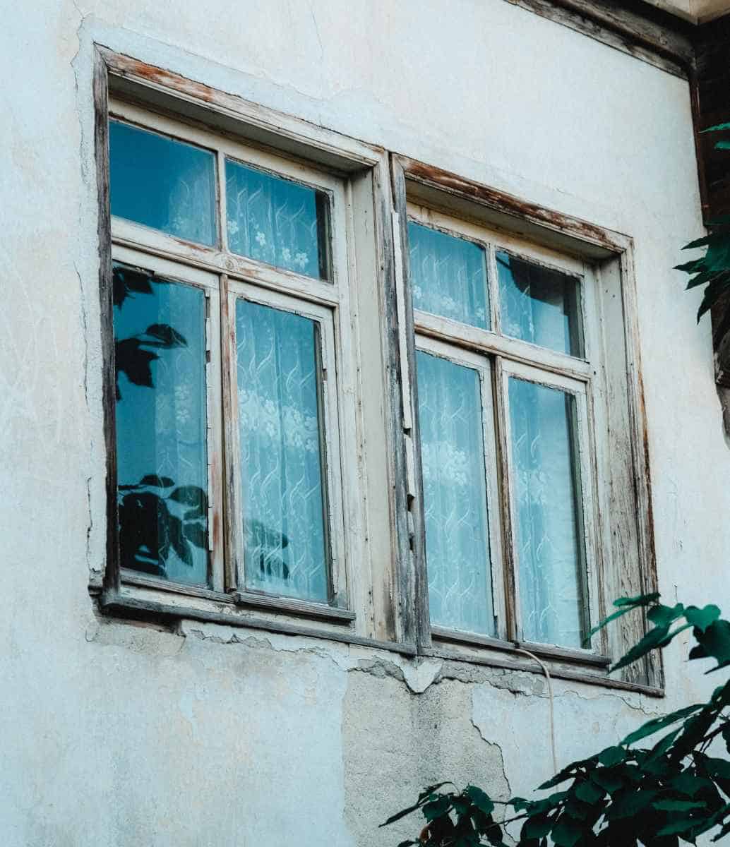 Ferestre vechi din lemn cu vopsea exfoliată și ramă degradată — tencuiala crăpată și deteriorată sub fereastră din cauza infiltrațiilor cauzate de lipsa protecției adecvate