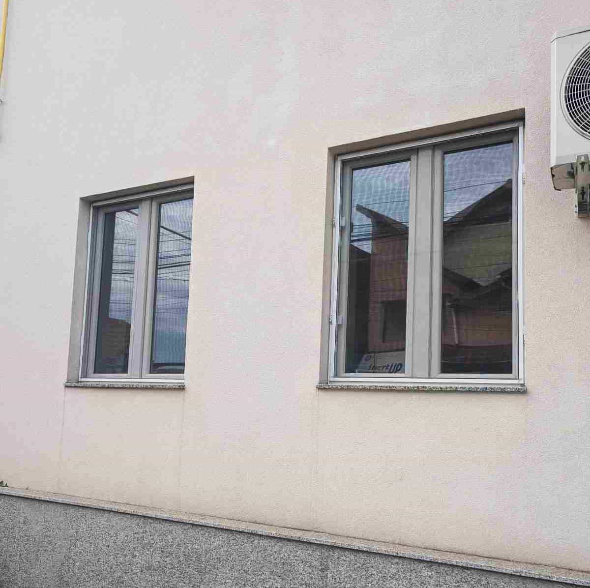 Glafuri exterioare din travertin pe o clădire cu tâmplărie PVC — urmele de scurgere pe fațadă sub fiecare fereastră indică lipsa soluțiilor laterale de protecție